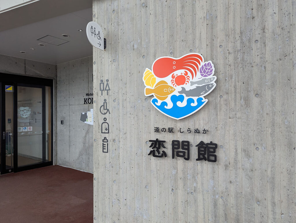コンクリート壁に設置された「道の駅しらぬか恋問館」のカラフルなロゴマーク。カニ・サケ・ホッキ貝など白糠の海産物が描かれている。左奥に自動ドアの入口が見える。